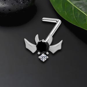 May include: Silver-toned nose ring featuring a bat-wing design with a large black gem center and a smaller, clear gem below. The L-shaped post is visible, designed for piercing. The jewelry is set against a dark background.