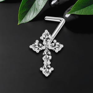 Può includere: Un piercing al naso argentato con un design a croce, ornato da pietre preziose chiare e scintillanti. La croce è composta da quattro pietre a forma di petalo e tre pietre aggiuntive sottostanti. Il piercing è posto su uno sfondo nero con foglie verdi.