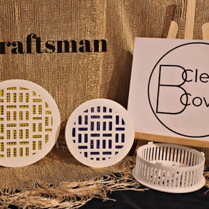 May include: Three white plastic drain covers with different patterns. The covers are round and have a grid pattern. The text "Clever Covers" is visible on a white sign behind the covers.
