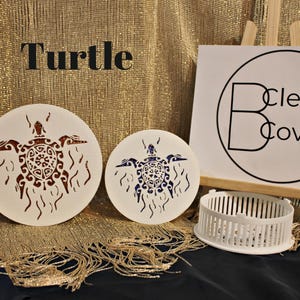 May include: Two white wooden round coasters with a turtle design. The turtle on the left coaster is brown and the turtle on the right coaster is blue. The coasters are on a gold background with the text "Turtle" and a white sign with the text "Clever Covers".