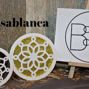 May include: Two white round decorative covers with intricate cut-out designs. One cover has a yellow background with a white design, and the other has a black background with a white design. The covers are labeled "Casablanca" and "Clever Covers".