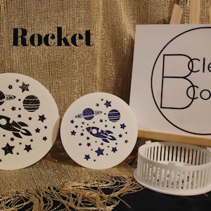 May include: Two white round stencils with black and blue designs of a rocket, planets, and stars. The text "Rocket" is above the stencils. The text "Clever Covers" is on a white sign behind the stencils.