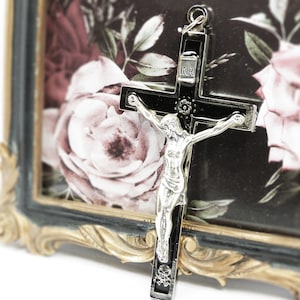 Könnte beinhalten: Ein silbernes Kruzifix mit einem schwarzen Kreuz und einer detaillierten Figur Jesu Christi. Das Kreuz hängt vor einem gerahmten Bild mit einem floralen Rosendesign. Der Rahmen hat ein schwarzes und goldenes, verziertes Design.