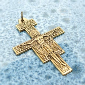 Könnte beinhalten: Ein goldfarbener Anhänger in Form eines Kreuzes mit einer detaillierten Darstellung von Jesus Christus am Kreuz. Das Kreuz hat aufwendige Schnitzereien und ist an einer kleinen Schlaufe oben aufgehängt. Der Hintergrund ist eine strukturierte blaue Oberfläche.