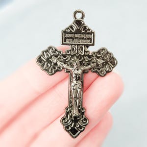 Puede incluir: Un colgante de crucifijo de metal oscuro con una figura detallada de Jesucristo en el centro. La cruz tiene bordes ornamentados y una placa rectangular en la parte superior con la inscripción JESUS NAZARENUS REX JUDAEORUM.