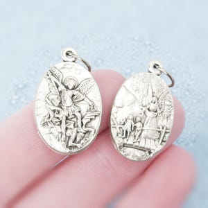 Op de afbeelding: Twee ovale hangers van zilverkleurig metaal met reliëf van religieuze afbeeldingen. De linkerhanger toont een engel boven een figuur, terwijl de rechterhanger een engel toont die over twee kinderen waakt bij een brug en een hek.