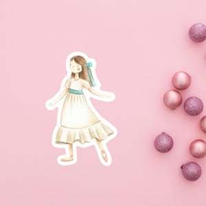 May include: A watercolor illustration of a girl in a white dress with a blue ribbon and sash. The image is on a pink background with several pink and glittery Christmas ornaments.