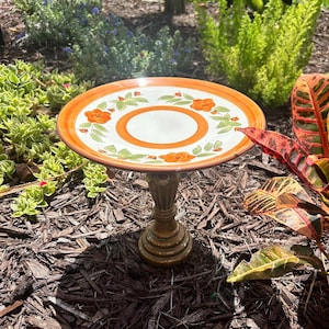 May include: A ceramic bird bath featuring a round, shallow basin with a white centre, an orange ring, and a wide orange rim. The basin is decorated with painted orange flowers and green leaves. It is supported by a textured, dark bronze-coloured pedestal base.
