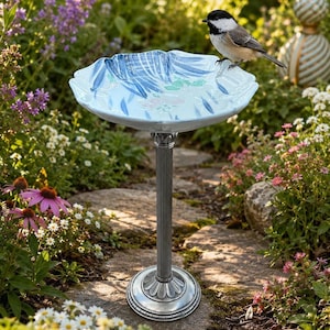 May include: A birdbath featuring a light blue ceramic basin with blue and green painted floral motifs, supported by a tall, fluted silver-toned metal pedestal base with a circular foundation, standing in a garden setting with a small bird.
