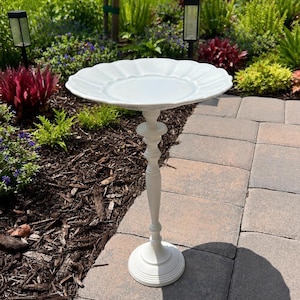 Puede incluir: Un baño para pájaros de color blanco con un cuenco ancho de bordes festoneados, sostenido por una base de pedestal delgada y decorativa. La estructura tiene un acabado en blanco mate sólido y una base circular escalonada para estabilidad.