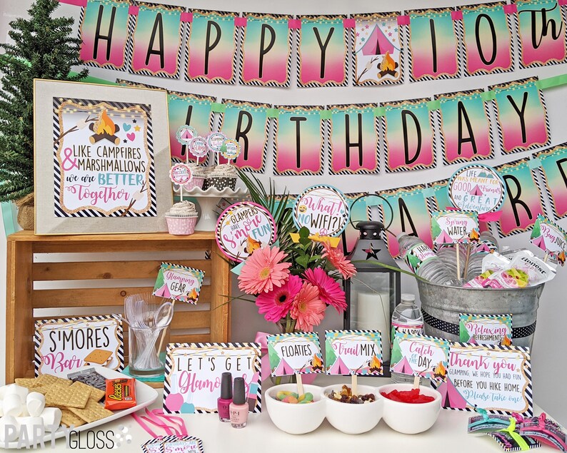 May include: A table setting for a camping themed birthday party. The table is decorated with a pink and green ombre banner that says "Happy 10th Birthday". There are also several signs with camping-themed sayings, such as "S'mores Bar" and "Let's Get Glamping".