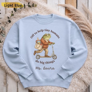 May include: A light blue crewneck sweatshirt featuring a graphic of a green frog wearing glasses and a brown jacket, riding a scooter while carrying a tote bag. The text reads: Off to help tiny humans do big things, followed by Ms. Laura.