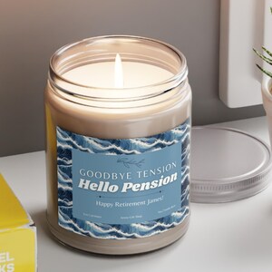 May include: A lit candle in a glass jar with a blue and white label that reads "Goodbye Tension, Hello Pension, Happy Retirement James!". The candle sits on a white surface.