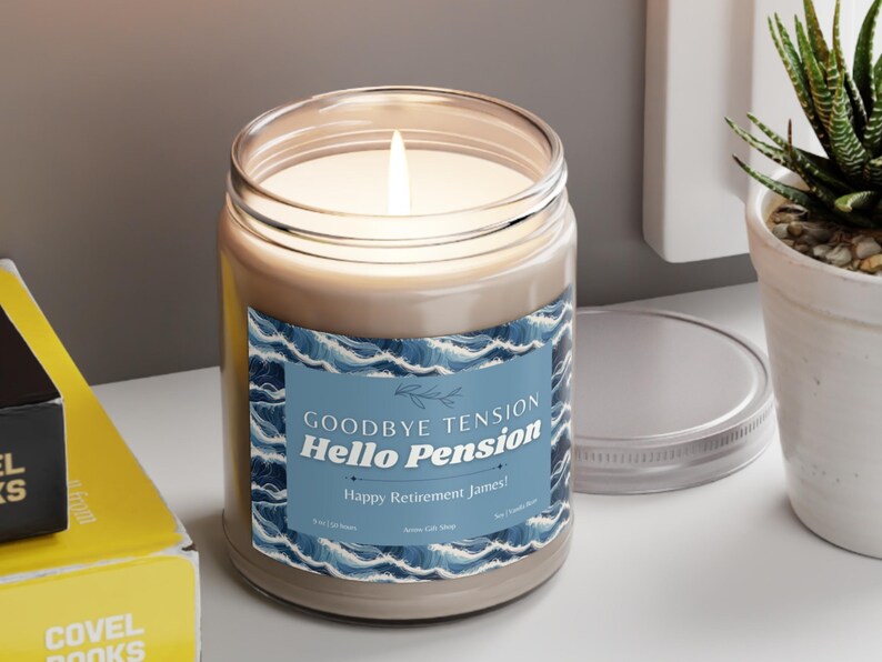 May include: A lit candle in a glass jar with a blue and white label that reads "Goodbye Tension, Hello Pension, Happy Retirement James!". The candle sits on a white surface.