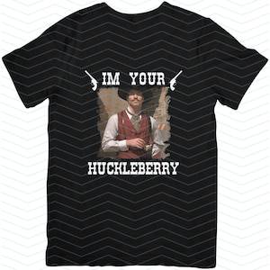 Puede incluir: Camiseta negra con un gráfico blanco de un hombre con un sombrero de vaquero y un chaleco sosteniendo un arma. El texto "I'M YOUR HUCKLEBERRY" está impreso en la camiseta.
