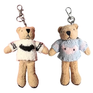 May include: Two brown teddy bear keychains wearing knitted sweaters. One bear is wearing a white sweater with a black design and the other is wearing a blue sweater with a pink design.
