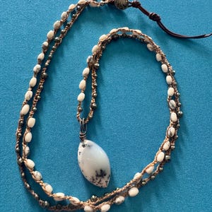 May include: A wrap necklace featuring a teardrop-shaped white stone pendant with dark dendritic inclusions, suspended from a brown cord woven with small white oval beads and metallic gray faceted beads, secured with a round button closure.