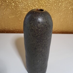 May include: A tall, cylindrical ceramic vase with a dark, mottled charcoal and brown finish. The surface has a textured, speckled appearance with a smooth, rounded top and a small, circular opening at the centre. The base is flat and stable.