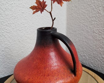 Puede incluir: Un jarrón de cerámica con cuerpo redondeado y asa negra. El jarrón tiene un esquema de color degradado, que va del rojo intenso en la base al negro en la parte superior. Contiene una ramita de hojas de arce artificiales con tonos naranja y marrón.