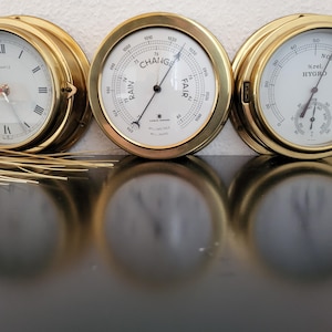 Könnte beinhalten: Drei nautische Instrumente mit Messingrahmen: eine Quarzuhr, ein Barometer und ein Hygrometer. Die Uhr hat römische Ziffern. Das Barometer zeigt "Change" und "Fair" an. Das Hygrometer zeigt "Normal" und %rel. HYGRO an. Alle haben weiße Zifferblätter.