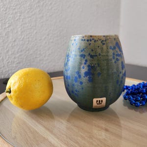 May include: A blue and green ceramic cup with a speckled glaze. The cup is sitting on a wooden surface with a lemon and blue beads.