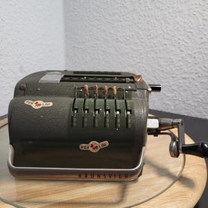 Op de afbeelding: Een olijfgroene Brunsviga mechanische rekenmachine met een slinger en cijferwielen. De rekenmachine heeft de tekst "PER LIM" en "BRUNSVIGA" erop. De rekenmachine staat op een houten oppervlak.