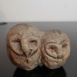 May include: A small, textured, tan-colored ceramic sculpture depicting two owl heads joined together. The owls have closed eyes, prominent beaks, and a rough, weathered surface finish that mimics the appearance of natural stone or clay.