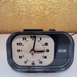 May include: A rectangular black alarm clock with a cream-coloured circular clock face featuring black Arabic numerals from 1 to 12. The clock has black hour and minute hands, a thin silver second hand, and the brand name ESGE printed in white.