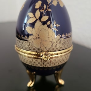 May include: A dark blue egg-shaped trinket box featuring gold floral patterns and a textured cream-colored band around the center. The box has a gold-toned metal hinge with a flower-shaped clasp and stands on three small, curved gold-colored legs.