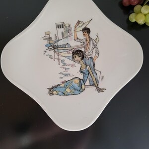 Peut inclure: Assiette décorative blanche en forme de losange avec une illustration colorée d'un couple près d'un front de mer. La femme porte une robe bleue et l'homme tient un chapeau. Des bateaux et des bâtiments sont en arrière-plan.