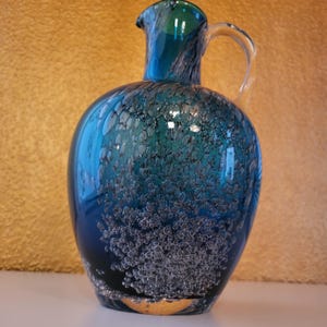 Può includere: Una brocca in vetro soffiato a mano caratterizzata da una sfumatura di colore blu e verde acqua intenso. Il corpo del recipiente è densamente riempito di numerose piccole bolle d'aria intrappolate nel vetro e presenta un manico curvo trasparente.