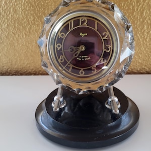 May include: A vintage Majak clock with a faceted crystal case and a dark burgundy face. The clock has gold numerals and hands, and rests on a black base. The text "Majak" and "11 jewels Made in USSR" are visible on the clock face.
