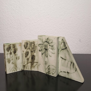 Könnte beinhalten: Vier cremefarbene Keramikplatten mit geprägten botanischen Designs. Die Platten zeigen Bilder von Trauben, Weinreben und einer Sonnenblume. Die Platten sind in einem Zickzackmuster auf einer dunklen Oberfläche angeordnet.