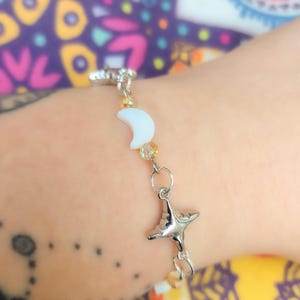 May include: Silver-toned bracelet featuring a crescent moon and a star charm. The bracelet includes small, light-colored beads and a clasp closure. The moon charm is white, and the star charm has a face. The bracelet is a celestial-themed accessory.