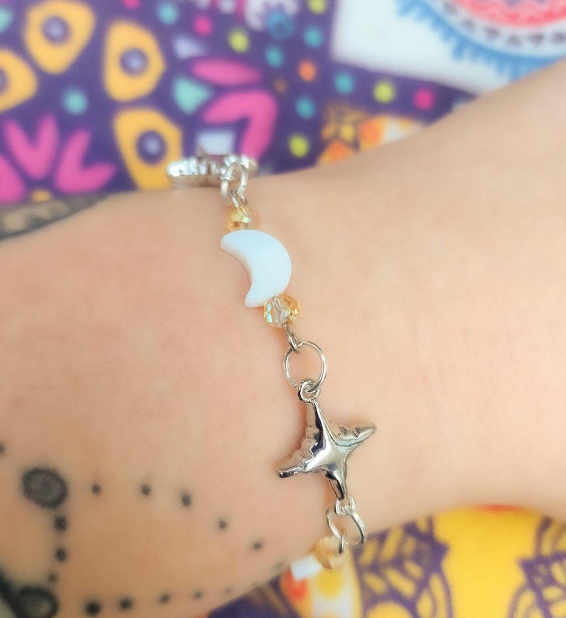 May include: Silver-toned bracelet featuring a crescent moon and a star charm. The bracelet includes small, light-colored beads and a clasp closure. The moon charm is white, and the star charm has a face. The bracelet is a celestial-themed accessory.