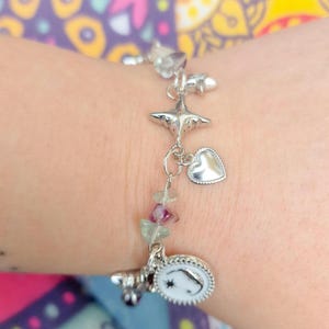 May include: Silver charm bracelet featuring star, heart, and moon-shaped charms. The bracelet includes a series of small, colorful gemstone beads. The bracelet is worn on a wrist.