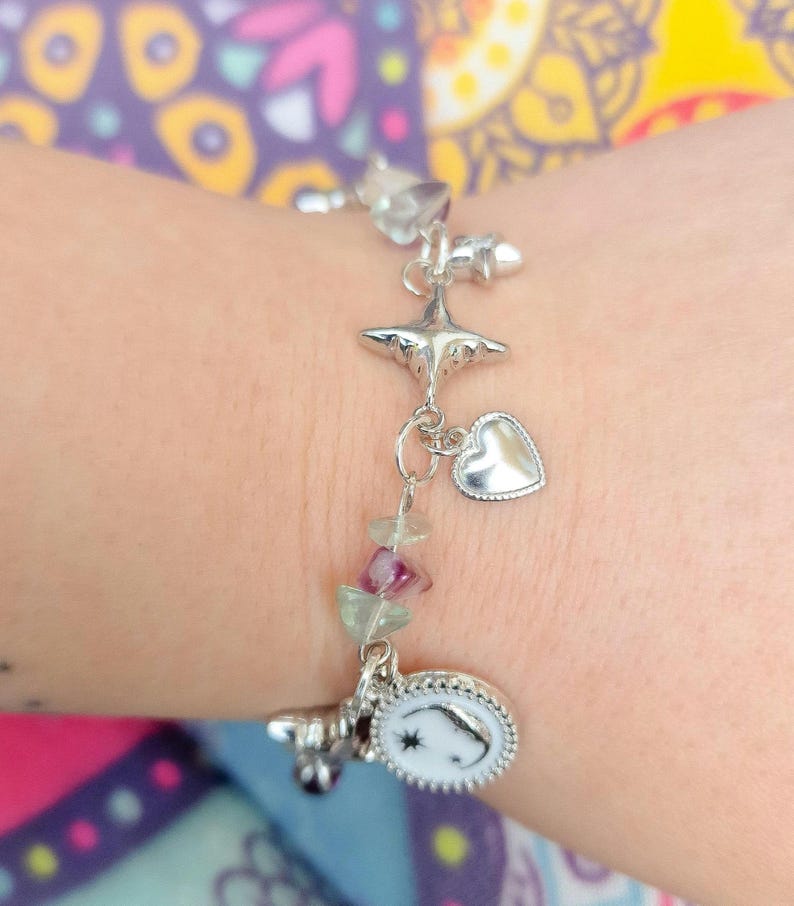 May include: Silver charm bracelet featuring star, heart, and moon-shaped charms. The bracelet includes a series of small, colorful gemstone beads. The bracelet is worn on a wrist.