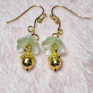 May include: A pair of dangle earrings featuring gold-tone metal acorn charms suspended from pale green translucent flower-shaped beads. The earrings are attached to gold-tone metal fishhook wires with a small spiral detail above the flower bead.