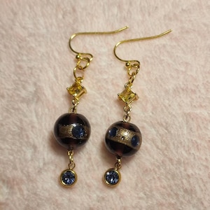 May include: A pair of gold-tone drop earrings featuring a small yellow square crystal, a larger dark purple spherical bead with a gold glitter band and blue accents, and a small round blue crystal dangling at the bottom on a gold hook wire.