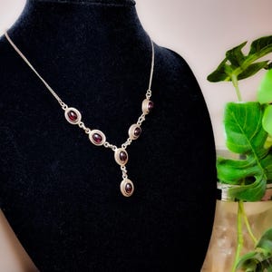 May include: A silver necklace featuring six oval-shaped dark red garnet stones set in metal bezels, arranged in a Y-shaped drop design. The necklace is displayed on a black bust, accompanied by a pair of matching oval garnet stud earrings.