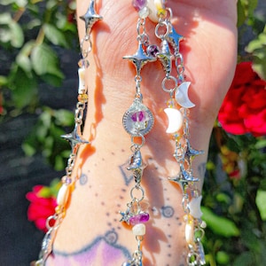May include: Silver-toned celestial-themed jewelry with star, moon, and crystal accents. The necklace features multiple strands with various charms and beads, including purple and white gemstones. The design incorporates a whimsical, bohemian aesthetic.