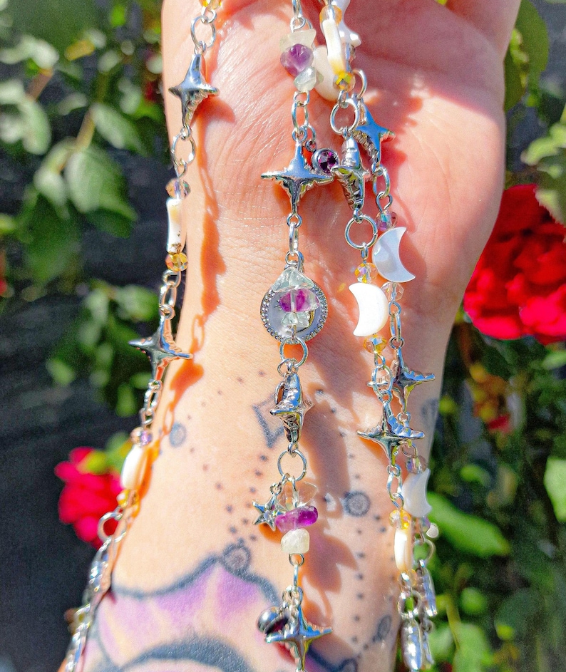 May include: Silver-toned celestial-themed jewelry with star, moon, and crystal accents. The necklace features multiple strands with various charms and beads, including purple and white gemstones. The design incorporates a whimsical, bohemian aesthetic.
