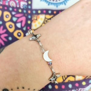 May include: Silver-toned bracelet featuring two stylized whale charms, a white crescent moon, and a small, iridescent crystal bead. The bracelet is strung with delicate chain links and clear beads. The bracelet is worn on a wrist.