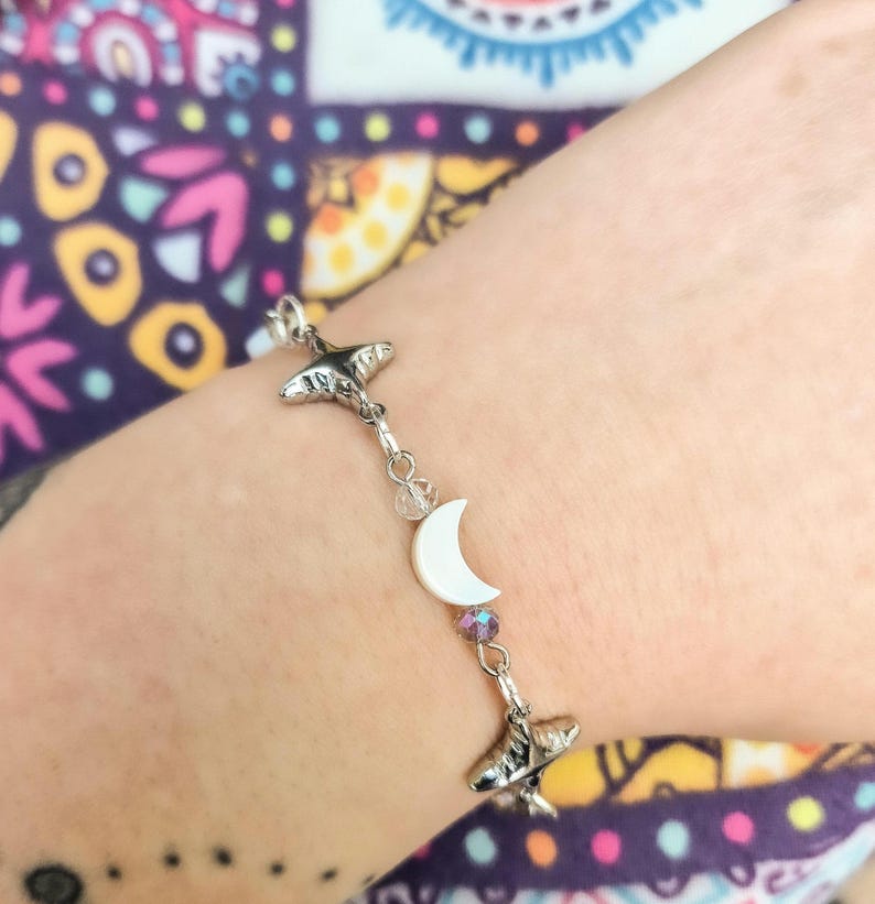 May include: Silver-toned bracelet featuring two stylized whale charms, a white crescent moon, and a small, iridescent crystal bead. The bracelet is strung with delicate chain links and clear beads. The bracelet is worn on a wrist.