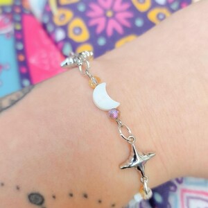 May include: A silver bracelet featuring a white crescent moon charm, star charms, and colorful beads. The bracelet is attached to a silver chain and is worn on a wrist.