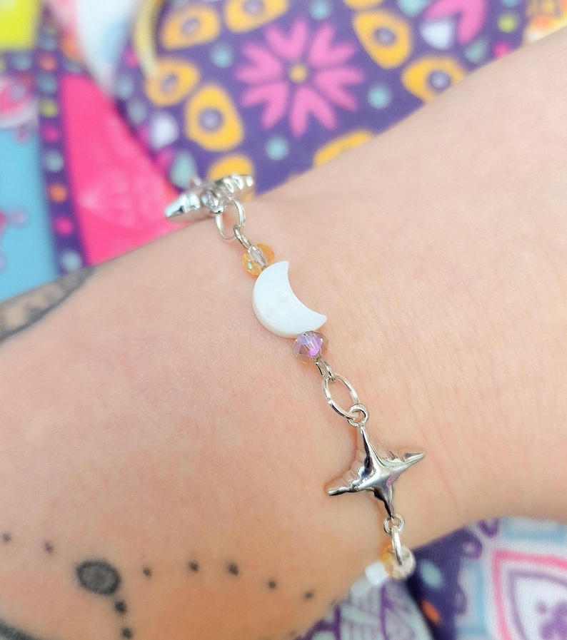 May include: A silver bracelet featuring a white crescent moon charm, star charms, and colorful beads. The bracelet is attached to a silver chain and is worn on a wrist.