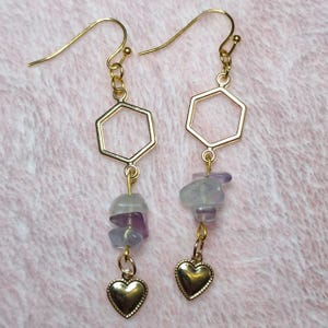 May include: A pair of gold-toned dangle earrings featuring a hexagonal frame, two irregular tumbled purple and white stone beads, and a small gold-toned heart charm at the base, suspended from gold-toned ear wires.