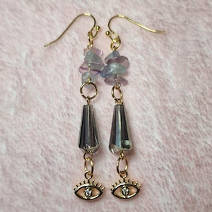 May include: A pair of gold-tone hook earrings featuring dangling purple and clear stone chips, a faceted gray crystal bead, and a gold-tone eye charm with a small clear rhinestone at the center of each eye, suspended from gold-tone metal wires.