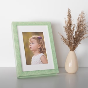Pode incluir: Uma moldura verde-clara com acabamento texturizado contendo a fotografia de uma criança pequena com um acessório de cabelo roxo. Ao lado da moldura, há um vaso de cerâmica bege com um ramo de ervas ornamentais secas e plumosas.