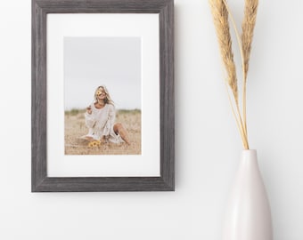 May include: A dark grey wooden picture frame with a white mount containing a photograph of a person in a white dress sitting in a field holding a yellow flower. Next to the frame is a white vase holding two tall, dried, tan-coloured stalks.